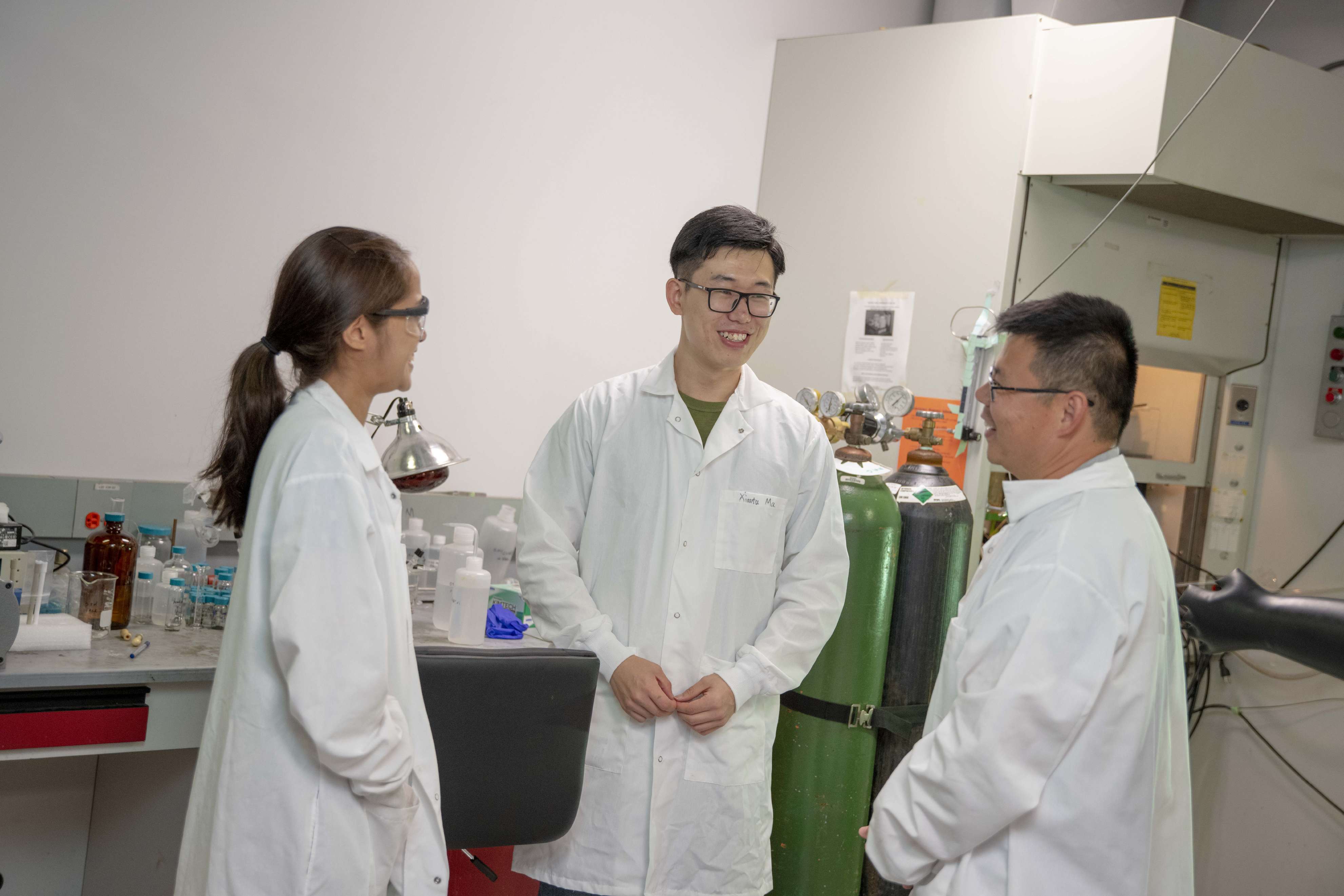 A man and woman in white lab coats laugh with Yan Wang, also in a white lab coat in a laboratory setting.