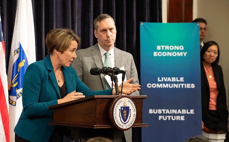 A woman wearing a green jacket speaks at a podium. A man in a gray suit is behind her.