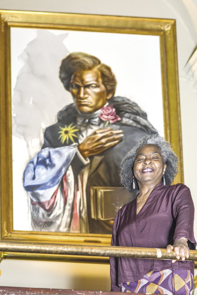 A woman wearing a purple shirt stands in front of a modern portrait of Frederick Douglass.