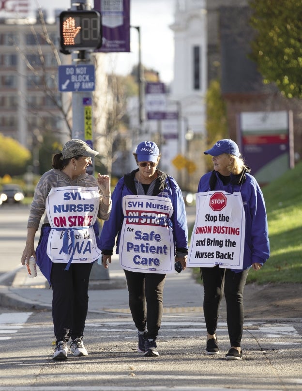 Three women wearing baseball caps walk across the street towards the camera wearing white signs around their necks with blue, red, and black lettering.