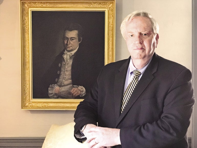 A man in a black suit and a yellow-striped tie stands in front of a portrait.