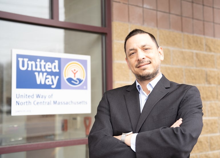 A man in a suit stands with his arms crossed in front of a window sign for the United Way