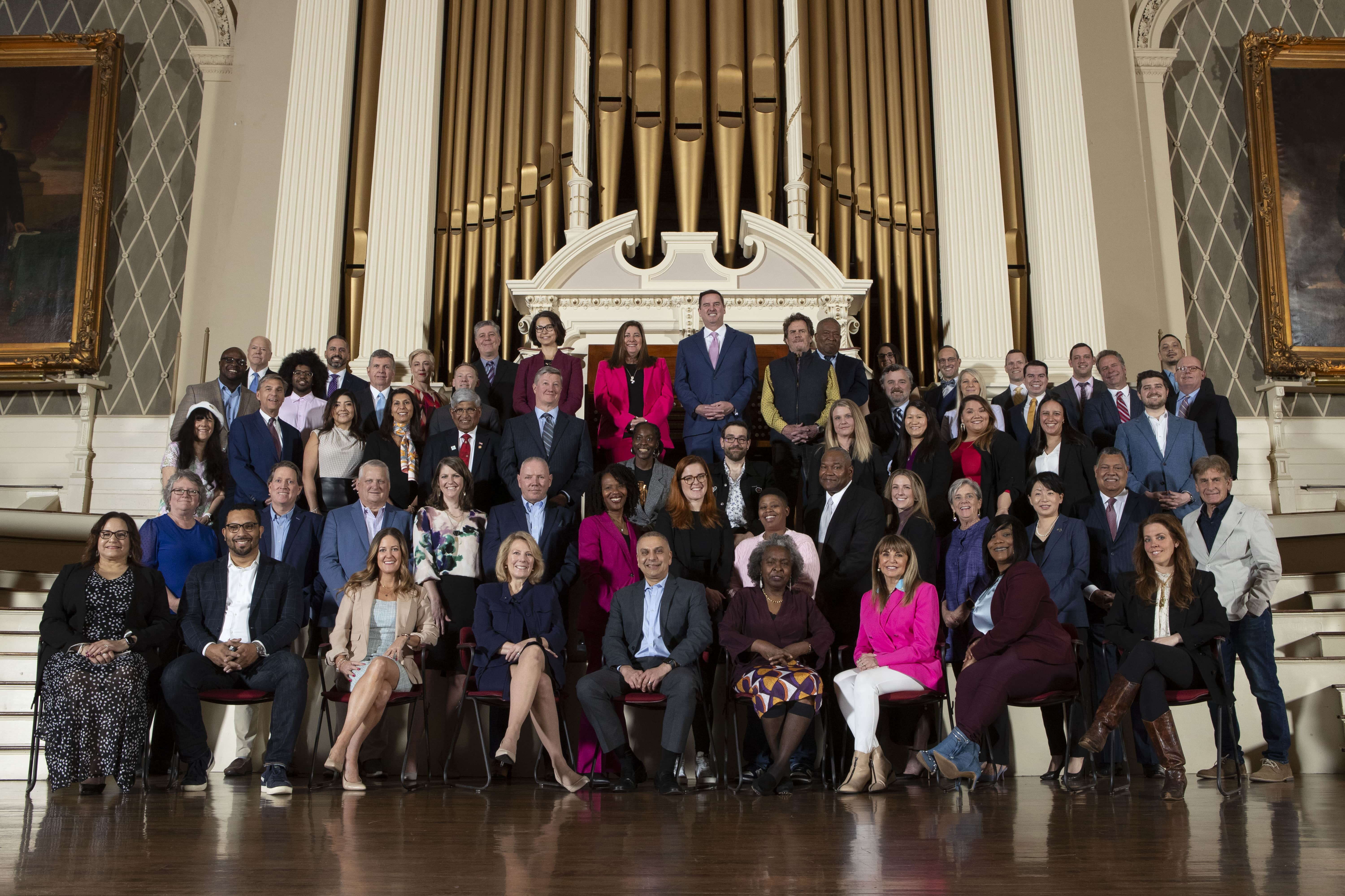 A group of 59 well-dressed professionals sit on a large stage together, looking at the camera.