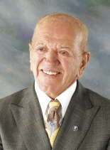 A headshot of a man wearing a grey suit jacket, white button down, and yellow, brown, and blue patterned tie