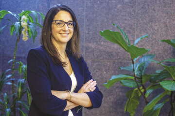 A woman with long brown hair wears glasses, a dark blue suit jacket, and white blouse standing with her arms crossed in front of green leafed plants.