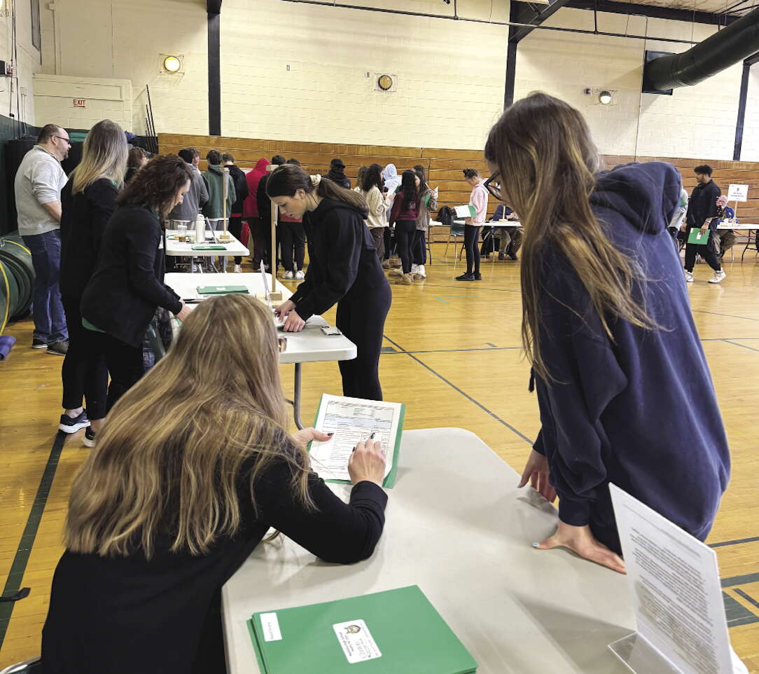 A woman with long blonde hair leans on a table showing papers to a student with long blonde hair wearing a purple sweatshirt in a school gymnasium with other adults and students at tables