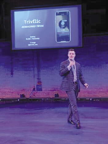 A man in a suit walks on a stage holding a microphone with a screen behind him
