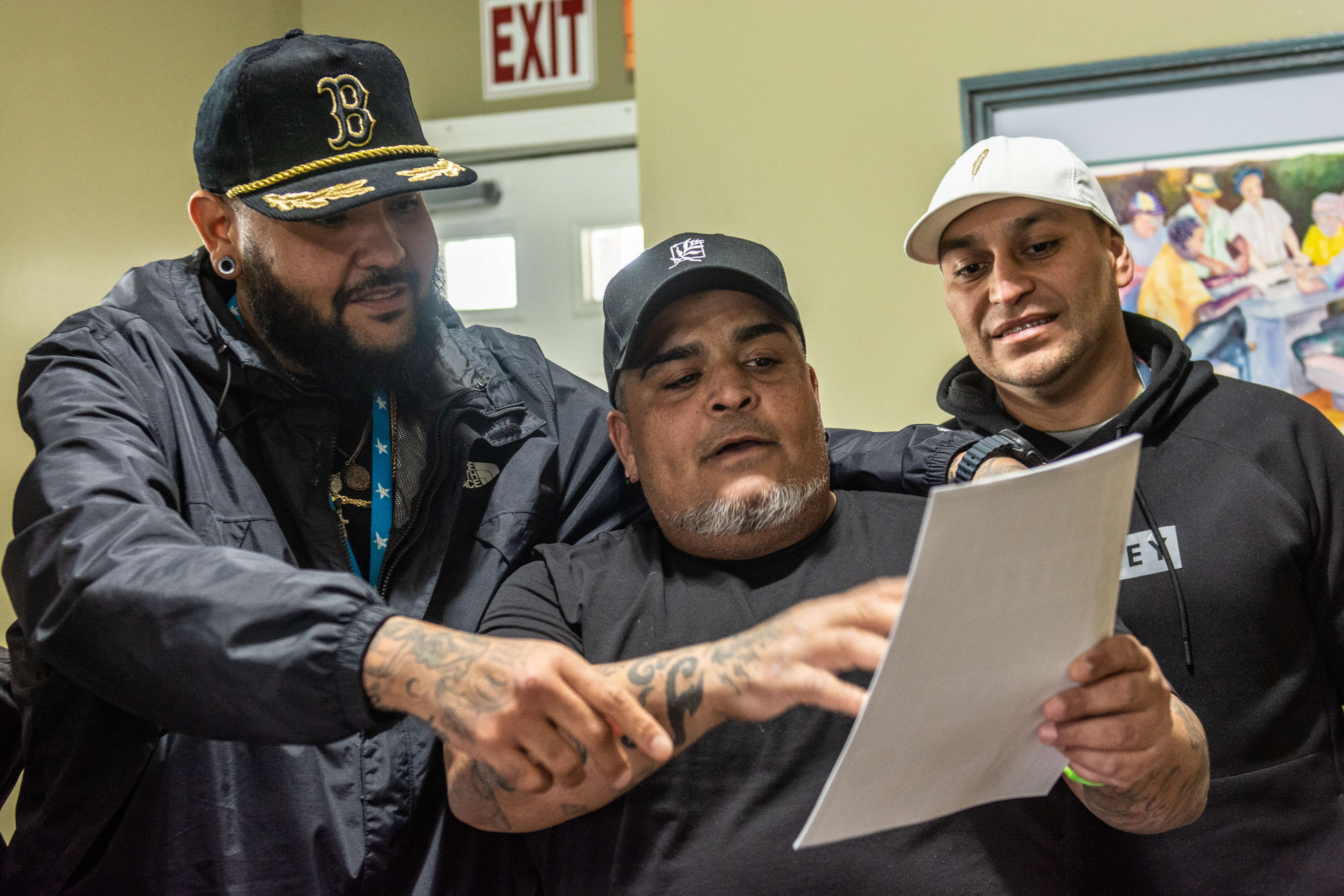 Three men wearing black clothes and baseball caps smile while looking at a piece of paper the middle one is holding.