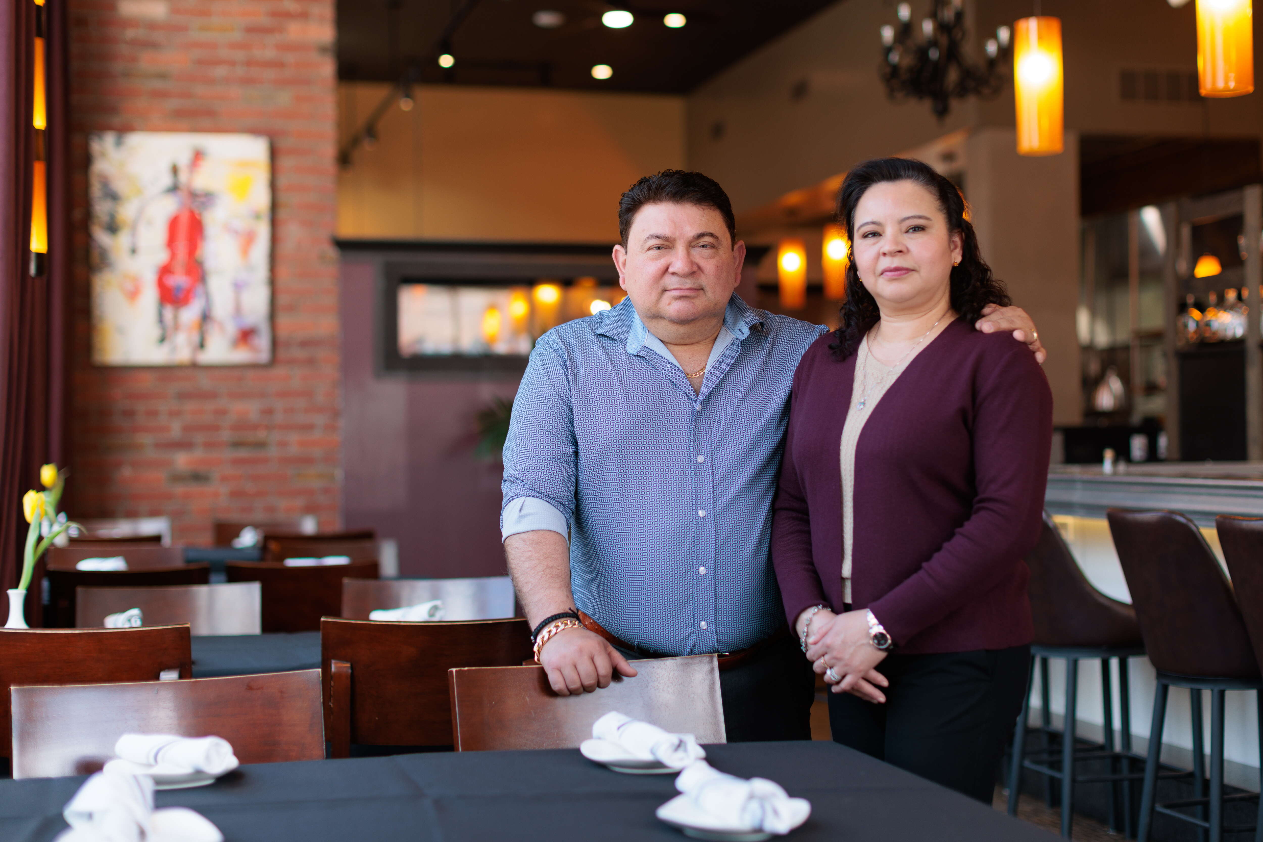 A man in a light blue checkered shirt and a woman in a maroon and beige sweater stand together in front of a restaurant table with chairs inside a restaurant.