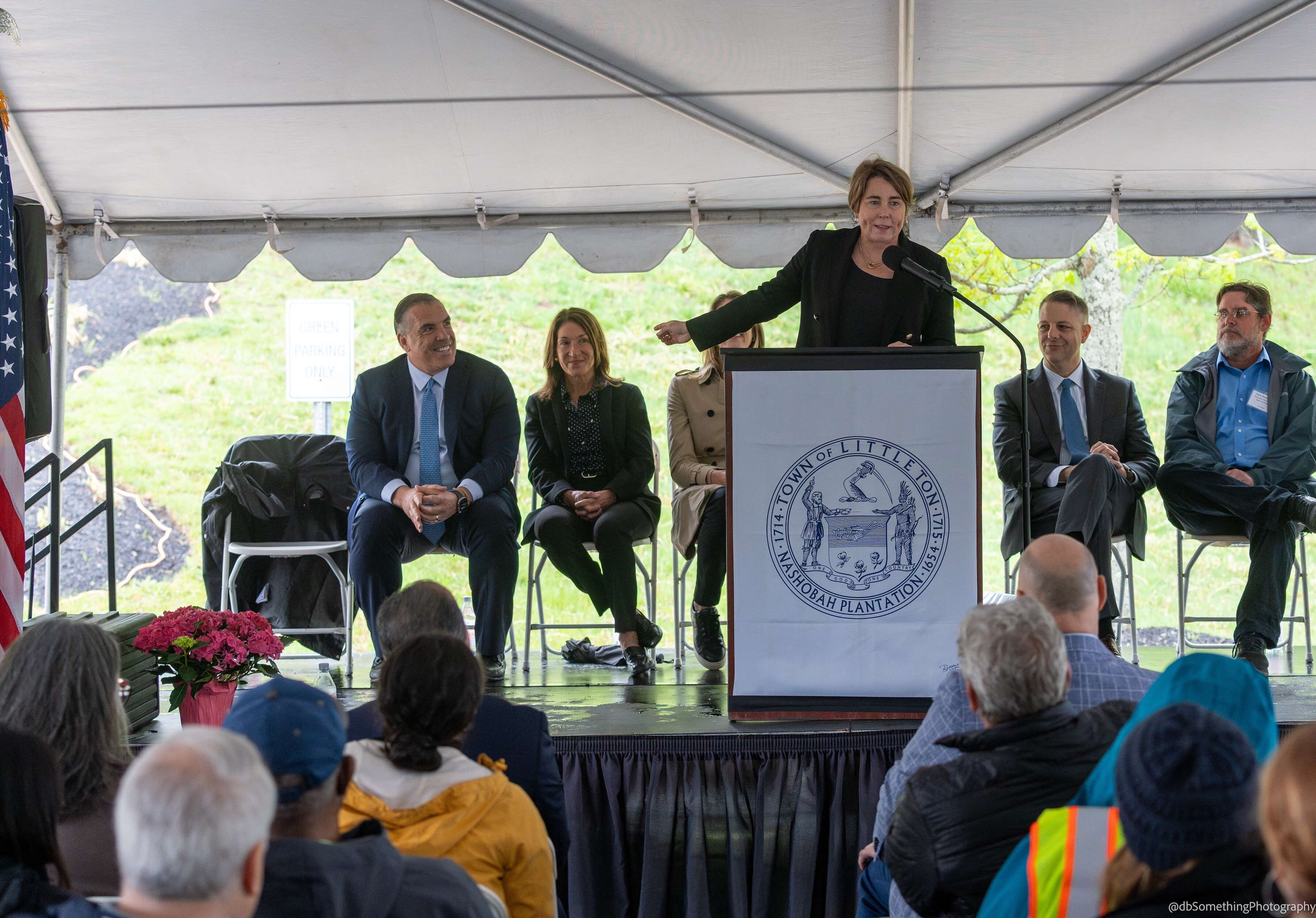 A woman at a podium on a stage with others sitting behind her points in the direction of a man on the stage