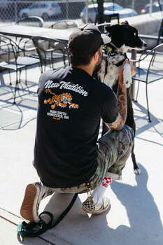 A man kneels in front of a black and white dog, holding it up outside.