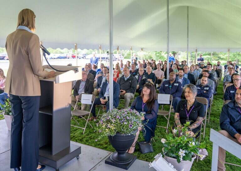 A woman speaks at a podium in front of a crowd seated under a tent outside.