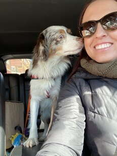 A woman wearing sunglasses and a black coat sits in a car with a dog.