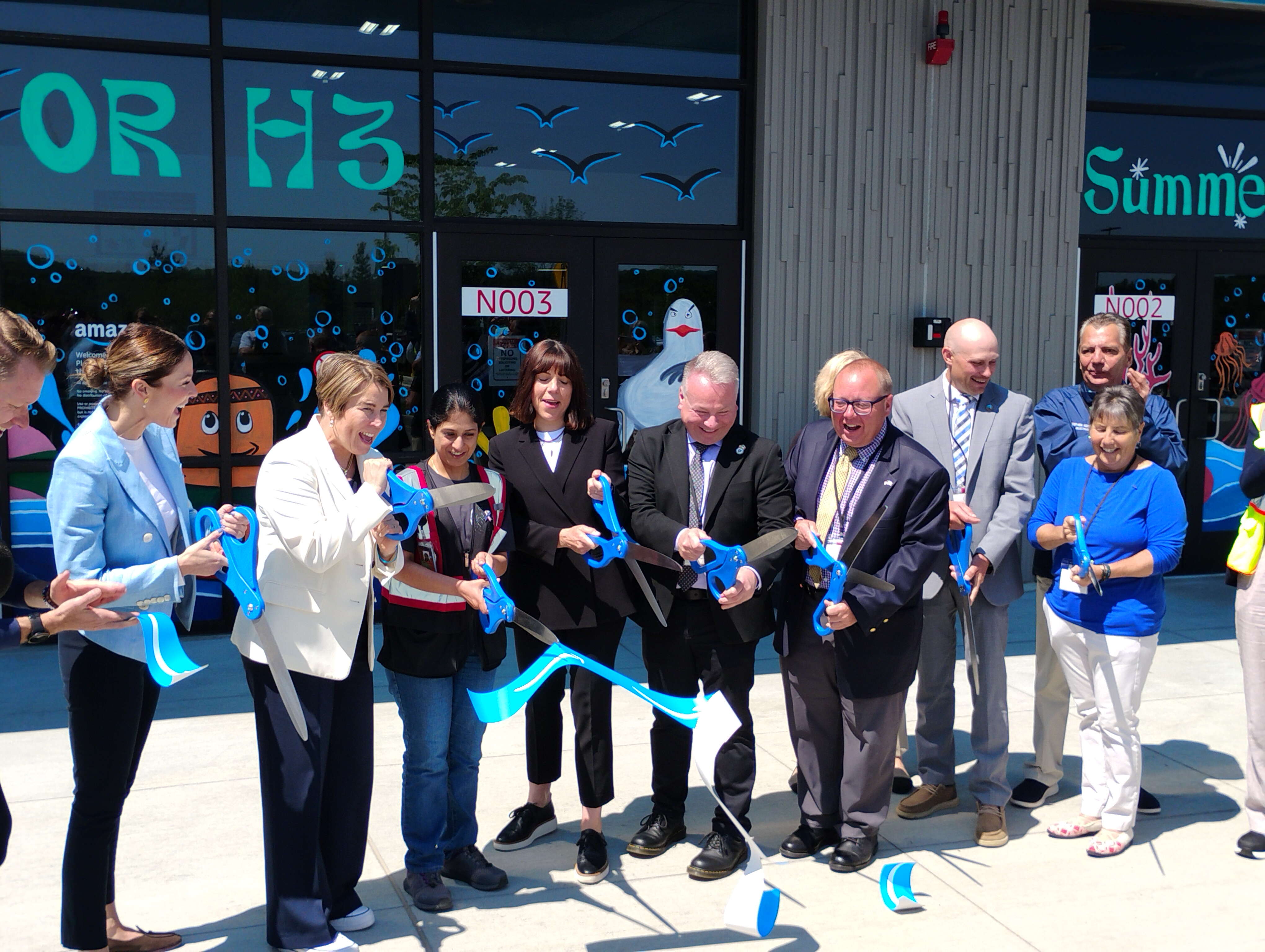 A group of people use ceremonial scissors to cut a blue ribbon in front of a building