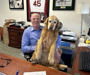 A man in a blue button down shits at a desk chair holding a large golden retriever on his lap.