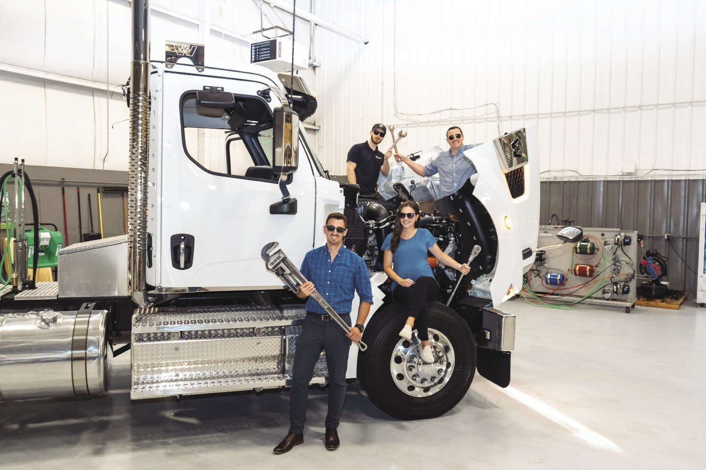 Four people holding wrenches while sitting on or around the open hood of a large white truck in a garage