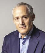 A man in a suit jacket sits in front of a grey background