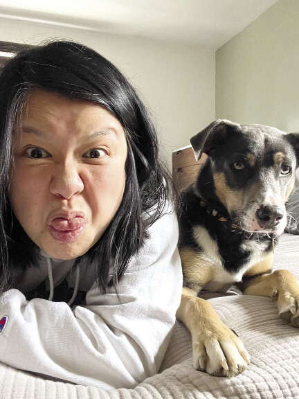A woman sits on a bed next to a dog while sticking her tongue out