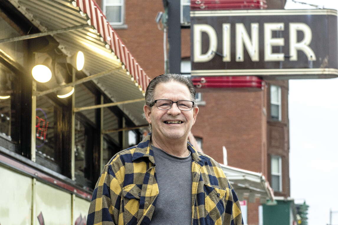 A man wears a grey shirt and a blue and yellow flannel shirt while standing in front of a diner with a sign above it reading "DINER".
