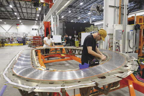 A man in a large manufacturing building works on a large circular magnet which he is standing in the middle of.
