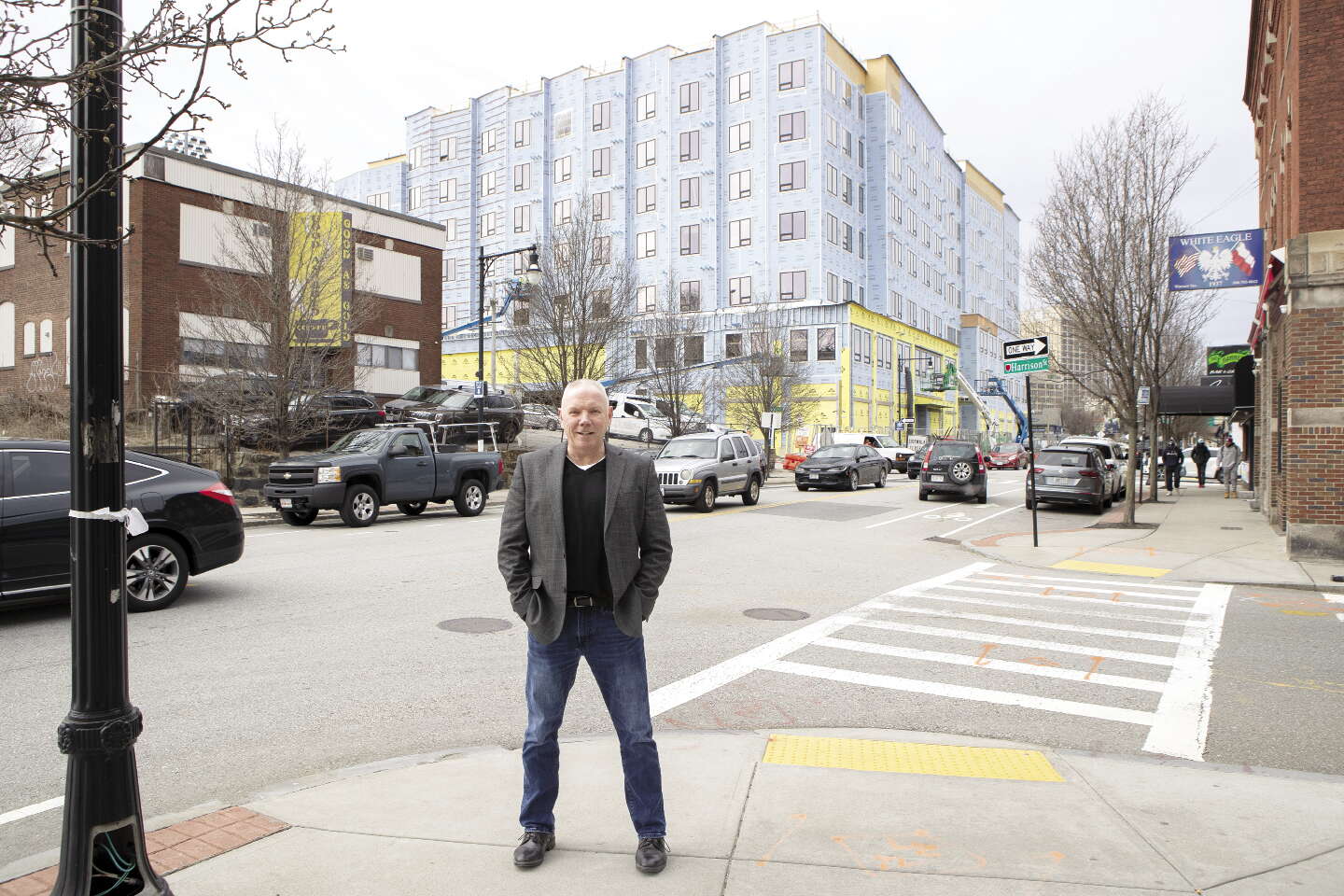 A man stands on a curb with his hands in his pocket facing the camera, standing across the street from a under-development apartment