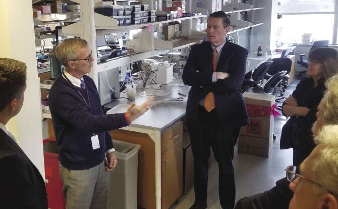 A man in a blue sweater speaks to a group inside a research space with packed shelves and papers spread amongst countertops