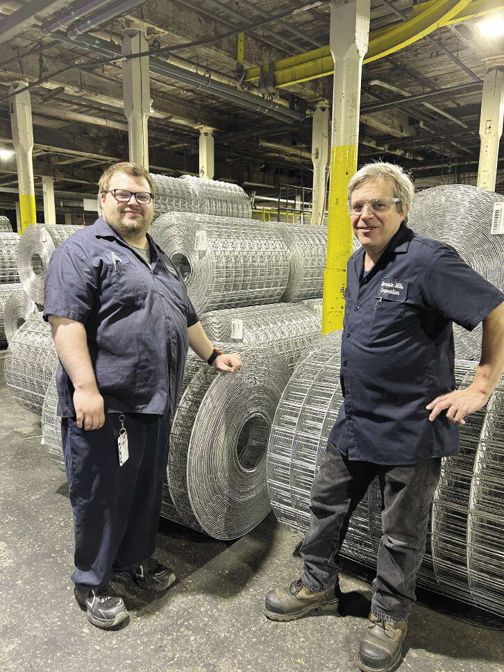 Two men wearing blue work shirts stand inside a warehouse and lean against large rolls of wire mesh.