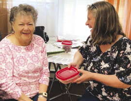 A woman with grey curly hair wearing a pink and whit top sits at a table laughing with a woman with shoulder length grey hair and a black and white print top.