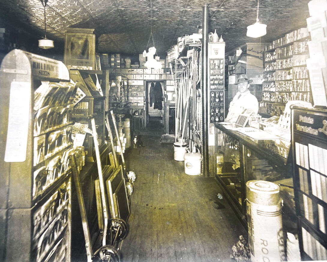 A black and white photo of the inside of a hardware store with a man in a fedora standing at the end of a glass counter on the right hand side, looking at the camera.