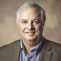 A man with gray hair wears a blue shirt and a gray jacket.