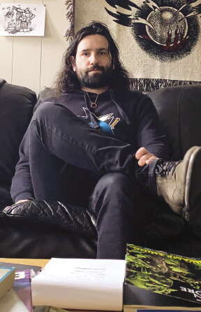 A man with long hair and a beard wears a sweatshirt, jeans, and boots sits on a black couch