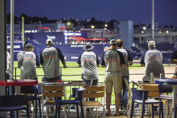 5 people facing away from the camera looking over a minor league baseball field. 