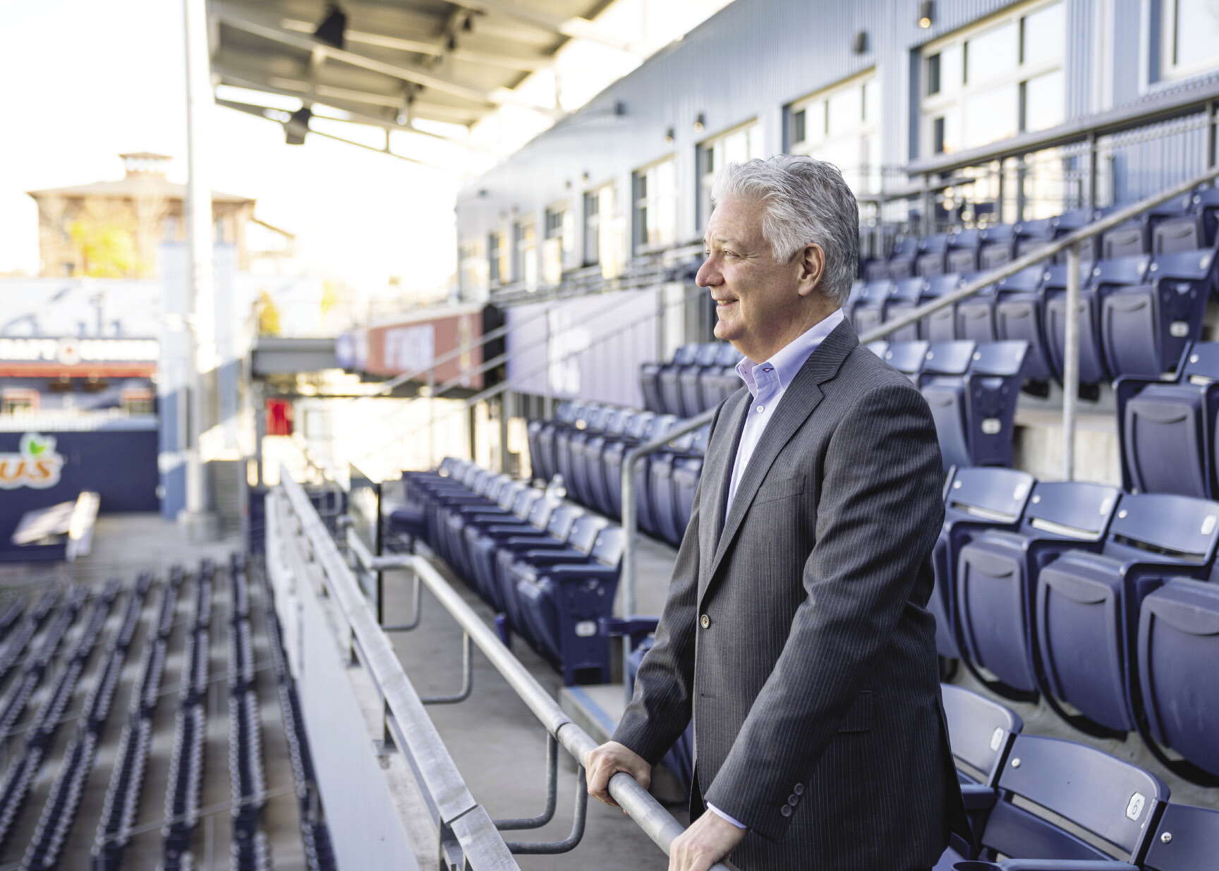 A man with gray hair and a pinstripe suit stands in a baseball stadium, looking outward.