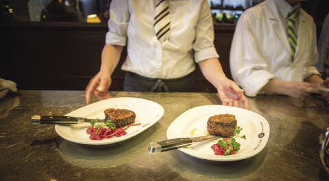 Waiters wearing white button downs and striped ties stand in front of a table with two white plates holding steak