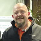 A man with a goatee wears an orange shirt and black jacket.