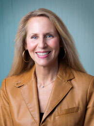 A woman with long blonde hair wears hoop earrings and a light brown leather jacket.