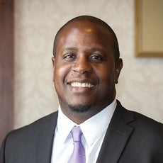 A man in a black suit jacket, white button down, and purple tie with small white dots smiles for the camera.