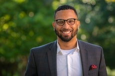 A man with dark hair, a beard, and black glasses wears a grey striped suit with a red pocket square and a white button down