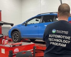 A blue car sits on a jack as a repair person tends to it. 