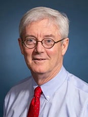 A headshot photo of a man with glasses, wearing a tie