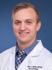 A man wearing a white lab coat and a blue shirt and tie smiles at the camera.