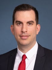 A man wearing a black suit with a white shirt and red tie looks at the camera.