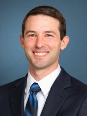 A man wearing a blue suit, white shirt, and blue-striped tie smiles at the camera.