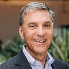 A man with salt-and-pepper hair wears a blue shirt and suit while smiling at the camera.
