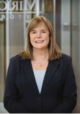 A woman with sandy blonde hair and bangs, a peark necklace, dark navy jacket and light blue shirt.