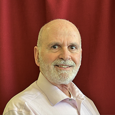 A man with a white beard and a light beige button down smiles for the camera.