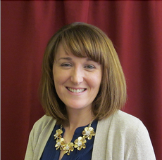 A woman with shoulder length hair and bangs wears a white cardigan, blue blouse, and gold flower necklace.