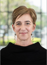 A woman with short hair wears a black mock-neck sweater and long earrings.
