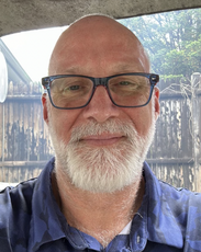 A man with glasses wears a blue patterned polo in front of a brown fence.