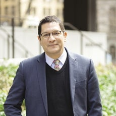Headshot of a man with dark brown hair wearing round glasses, a dark grey suit jacket, black sweater, white button down, and light pink and green tie